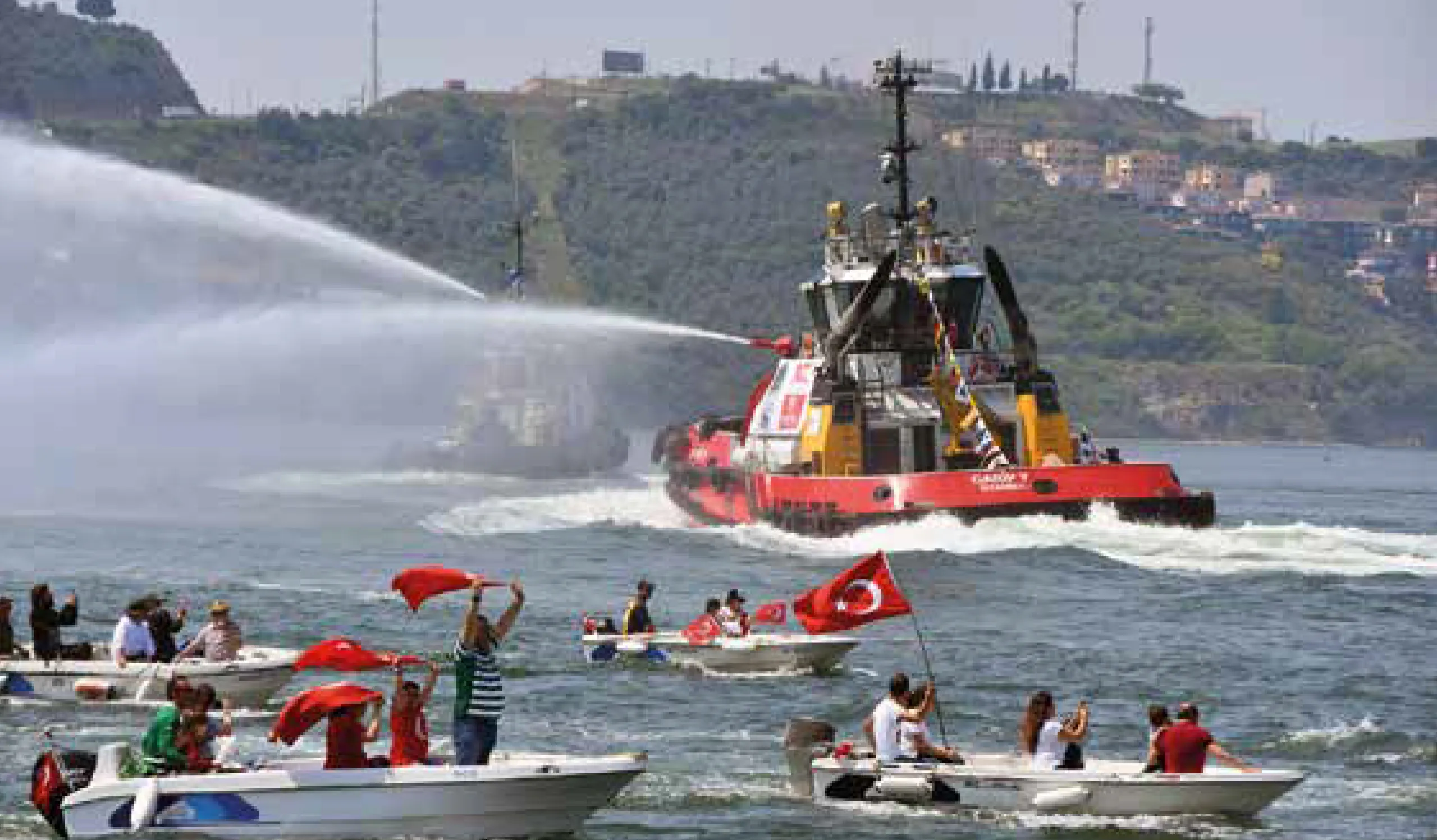 Boats adorned with Turkish flags celebrating the maritime festival, accompanied by a water-spraying tugboat.